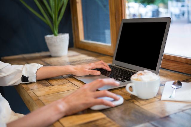 Young woman using laptop in a cafeteria, hands typing on keyboard. Coffee cup and spoon on wooden table, plant in background. Ideal for illustrating remote work, freelancing, modern workspaces, and casual work environments.