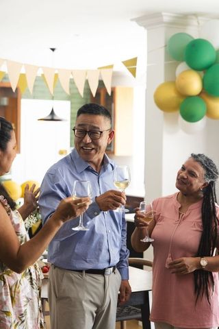 Mature friends toasting with white wine and colorful balloons in home kitchen celebration