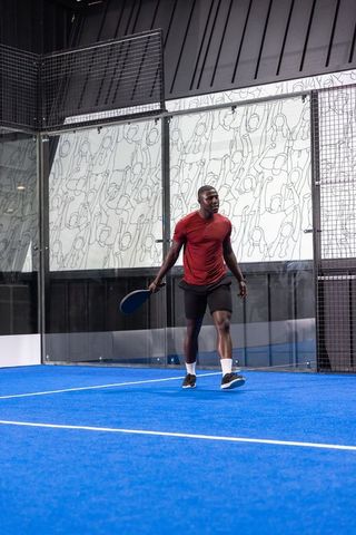 African american athlete playing padel on vibrant blue court with glass walls and mural