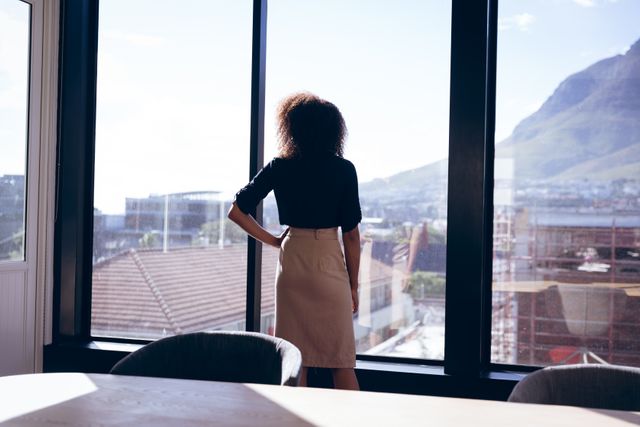 This image shows a businesswoman taking a break and looking out of a large office window, admiring the cityscape. It can be used to illustrate themes of professional life, career contemplation, urban work environments, and moments of inspiration or reflection in business settings.