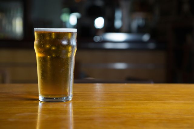 Close-up shot of a beer glass filled with amber beer placed on a wooden counter in a bar. The background is blurred, highlighting the drink in focus. Ideal for use in advertisements and promotions for bars, pubs, and breweries, or in articles relating to nightlife, socializing, and beverages.