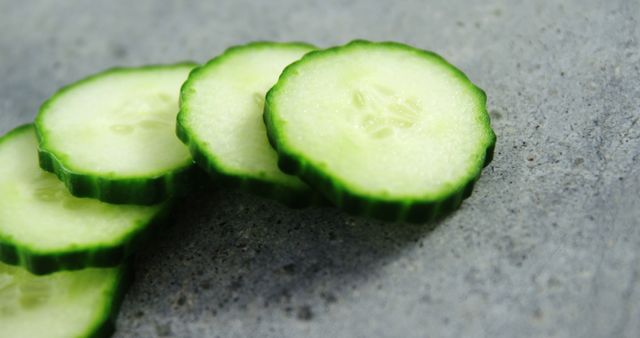 Sliced cucumbers are arranged on a textured grey surface, showcasing their fresh, green interior. Cucumbers like these are commonly used in culinary dishes for their crisp texture and refreshing taste.