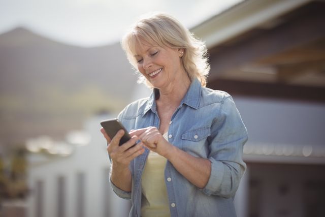 Senior woman smiling while using her mobile phone outdoors on a sunny day. She is dressed in casual clothing, including a denim shirt. This image can be used for themes related to technology use among the elderly, outdoor activities, modern communication, and happy lifestyles.