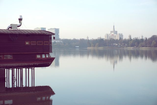 A tranquil setting featuring a wooden lakeside cabin on stilts with a distant city skyline. The calm waters reflect the serene environment, making it perfect for use in travel blogs, destination marketing, advertisements, or any media depicting tranquility amidst urban life.