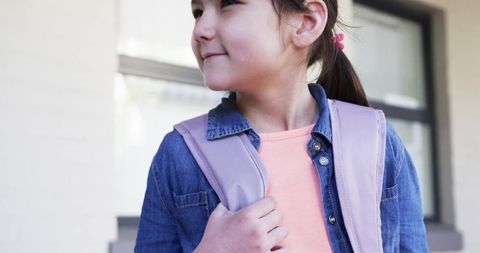 Young girl wearing denim jacket holding backpack strap and looking left before school