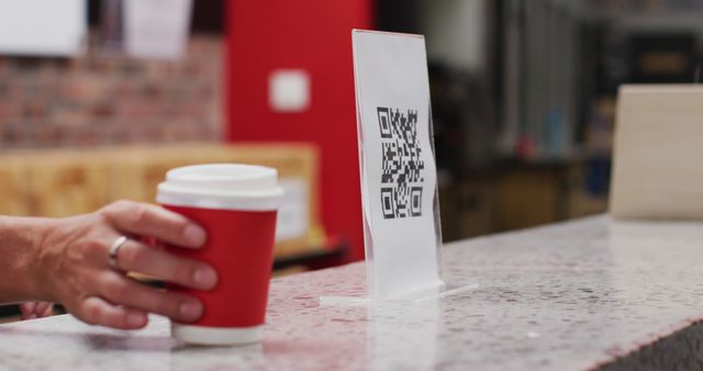 Hand holding a red cup is next to a printed QR code in a modern café. The QR code suggests the implementation of digital payment and contactless transactions. This moment captures the intersection of technology and everyday life, perfect for themes related to coffee culture, modern technology, and digital convenience. Suitable for articles or advertisements emphasizing digital payments in cafés or promoting contactless experiences.