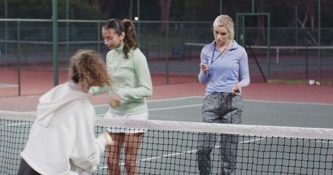 Female coach leading diverse players in nighttime tennis practice under court lights
