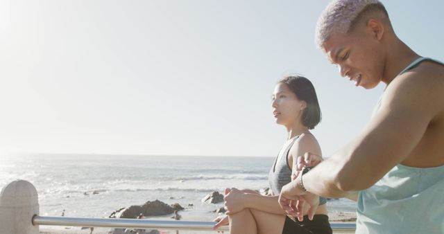 Fit couple takes a break during their morning workout by the ocean. Man checks fitness tracker while woman stretches, highlighting a modern, healthy, and active lifestyle. Perfect for use in fitness and wellness promotions, healthy living articles, and outdoor exercise inspirations.