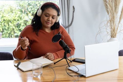 Asian woman recording podcast at home studio with laptop, microphone, taking notes