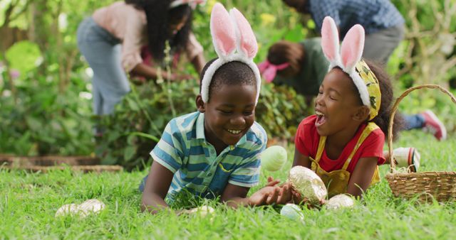 Two children lie on grass wearing bunny ears, collecting decorated Easter eggs and smiling with joy. The background shows adults participating in the activity. Perfect for illustrating family Easter celebrations, springtime traditions, and outdoor fun.