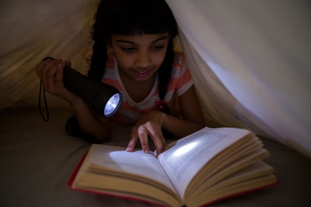 Girl holding flashlight while reading novel under blanket at home