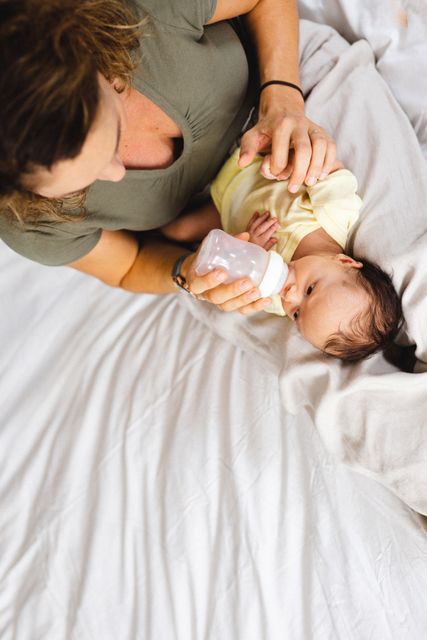 High angle view of a mid adult Caucasian mother feeding her newborn baby with a bottle while lying on a bed at home. This image can be used for parenting blogs, family care websites, or advertisements related to baby products and maternal care.