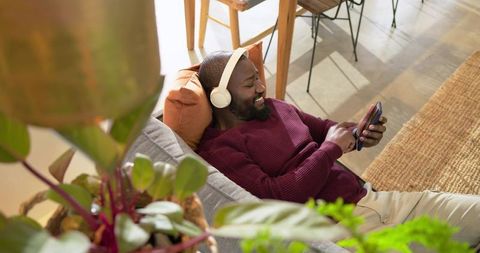 African american man relaxing on sofa with headphones and smartphone among indoor plants