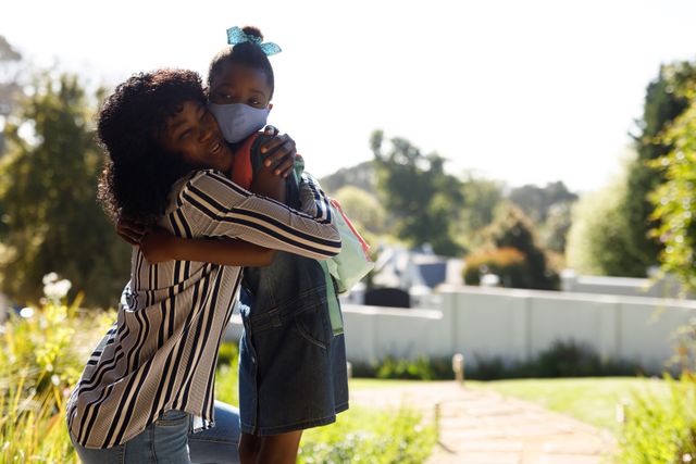 Close up of an african-american mother hugging with her daughter on the front porch. her daughter is wearing a facemask and a backpack from school