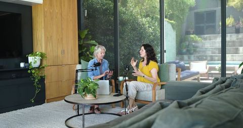 Mature women recording podcast in modern sunlit living room with microphones and laptop