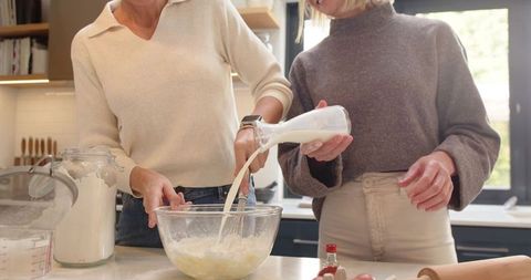 Two women whisking batter and pouring milk in sunlit kitchen for cozy home baking moment