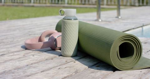 Olive-green yoga mat leaning on pool deck with textured bottle and pink headphones