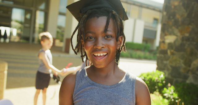Smiling African American child wearing graduation cap in sunny outdoor schoolyard. This image can be used to represent educational milestones, academic achievements, youth, and celebrations. Suitable for articles and advertisements related to education, back-to-school promotions, and children's educational programs.