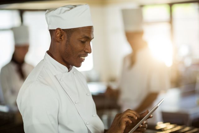 Chef in white uniform using digital tablet in a modern commercial kitchen. Perfect for restaurant or culinary industry websites, technology integration in kitchens, and professional chef profiles.