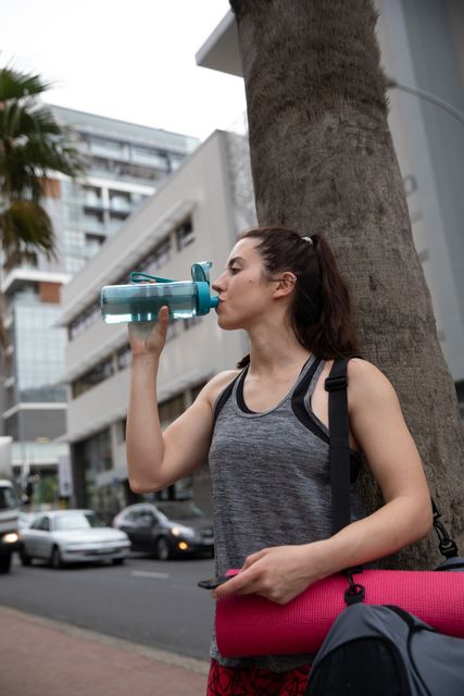  A fit Caucasian woman walking in the street on her way to fitness training, carrying a sports bag and a yoga mat, holding a bottle of water and drinking