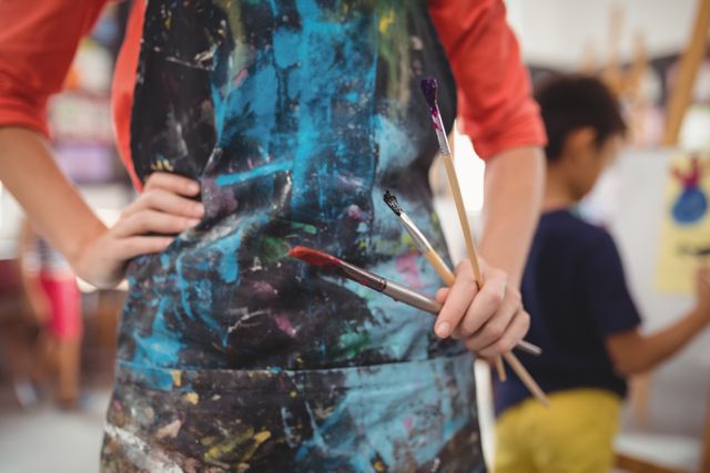 Art teacher holding paintbrushes in a colorful apron, with a child painting in the background. Ideal for educational content, art class promotions, creativity workshops, and school-related materials.