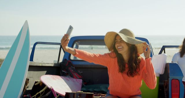 Young biracial woman takes a selfie at the beach, with copy space. She's capturing memories in a vibrant outdoor setting with surfboards.