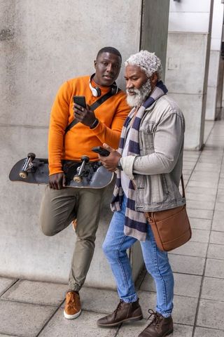 Urban multigenerational duo checking smartphones with skateboard on concrete corridor
