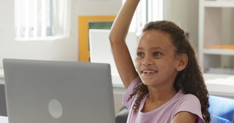 African american girl raising hand using laptop in bright classroom smiling