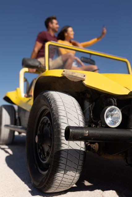 Happy caucasian couple sitting on beach buggy in the sun taking selfie, focus on foreground. beach stop off on romantic summer holiday road trip.