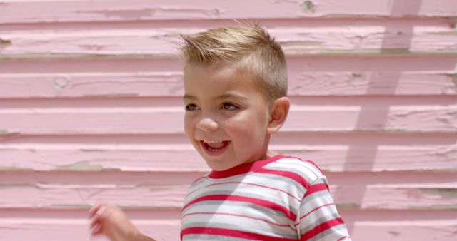 A joyful boy in a striped shirt enjoys playing outdoors on a sunny day. Bright pink wooden background adds a whimsical touch. Perfect for use in advertisements, children's clothing catalogs, or family lifestyle promotions.