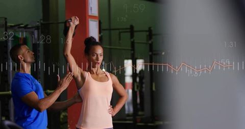 Trainer spotting woman performing kettlebell overhead press with performance data overlay