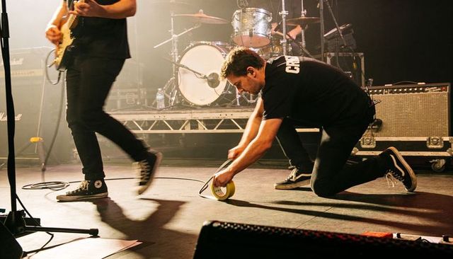 Crew member kneeling taping cable on live concert stage while guitarist walks by