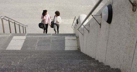 Two women walking up urban stairs carrying duffel and coffee cup, casual city friendship