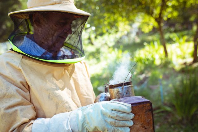 Caucasian senior man wearing beekeeper uniform preparing smoke to calm bees. beekeeping, apiary and honey production concept.