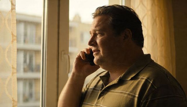 Middle-aged man talking on smartphone by apartment window with sheer curtains in afternoon light