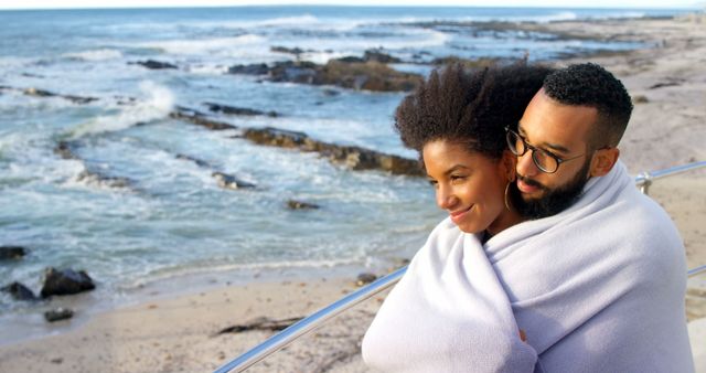 Romantic diverse couple with blankets on backs standing, smiling and embracing on sunny beach. Summer, vacation, romance, love, relationship, free time and lifestyle, unaltered.