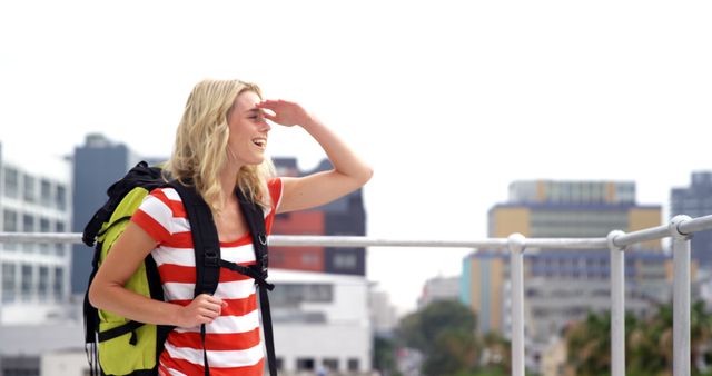 Blonde woman wearing striped shirt and backpack joyfully exploring urban area. Ideal for travel blogs, city exploration themes, adventure advertisements, and content about independent and carefree travel.