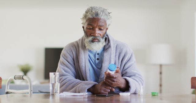 Elderly man with gray hair and beard carefully reading prescription bottle while sitting at table in home environment. Man is wearing comfortable robe and has glass of water next to him, emphasizing self-care and daily medication routine. Image can be used in healthcare, wellness, aging, patient education, and daily life contexts.