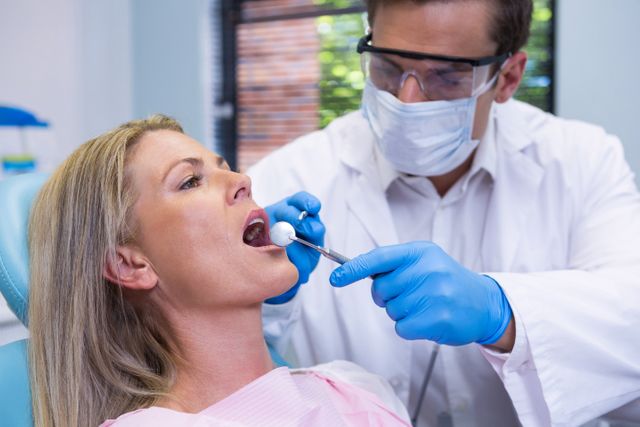 Dentist wearing surgical mask and gloves examining female patient in dental clinic. Useful for illustrating dental care, oral health, medical procedures, and healthcare services. Ideal for dental clinic websites, healthcare brochures, and educational materials on dental hygiene.