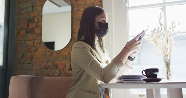 Woman in business casual attire wearing a protective mask sitting at a cafe table, using a smartphone while a cup of coffee sits in front. Sunlight coming through window. Ideal for concepts related to safe social interactions, working remotely, new normal, or casual work environment visuals.