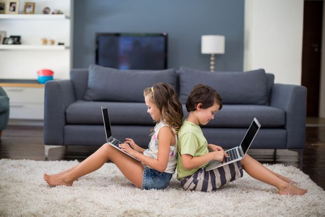 Smiling siblings sitting back to back and using laptop in living room at home