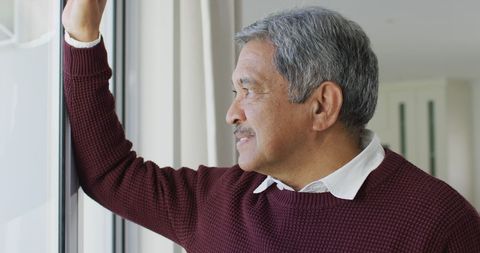 This image depicts a senior man with gray hair smiling thoughtfully while gazing out of a window in a relaxed home environment, suggesting contemplation and positivity. Useful for illustrating articles or advertisements related to retirement planning, mental health, daily life, senior living solutions, or personal reflection stories.