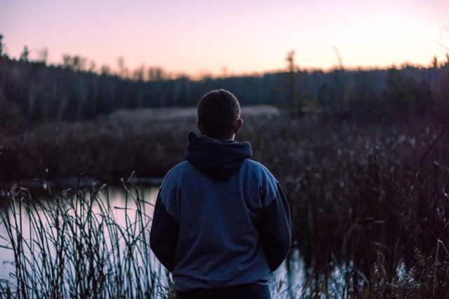 A person is standing by a forest lake during sunset, creating a peaceful and reflective scene. The individual is wearing a hoodie and is silhouetted against the serene natural backdrop of reeds and water. This can be used for themes of solitude, reflection, nature appreciation, tranquility, and outdoor activities.