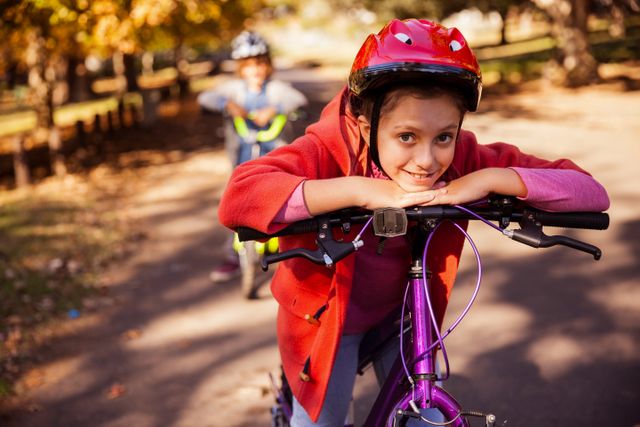 Young girl wearing helmet and red jacket leaning on mountain bike in park during autumn. Background shows another child with bike. Ideal for promoting outdoor activities, children's safety, and family recreation.