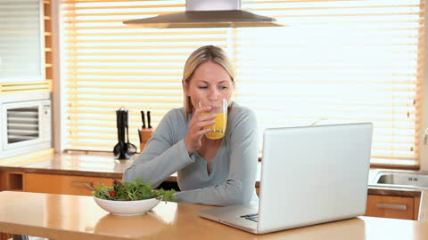 Woman Enjoying Orange Juice While Working on Laptop in Kitchen
