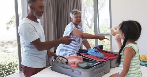 A biracial couple smiling while assisting their young daughter stuffing a suitcase, teaching her about responsibility and teamwork. Ideal for promoting family holidays, social interactions, education through family activities, travel preparations, or parenting articles.