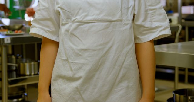 Professional chef dressed in white chef's uniform standing in a commercial kitchen. Surrounding environment filled with stainless steel kitchen equipment and utensils. Ideal for use in articles about culinary professions, commercial kitchens, restaurant operations, cooking tutorials, and hospitality industry visuals.