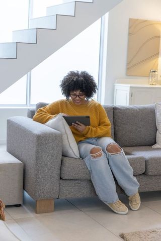 African american woman sitting on gray sofa using tablet in bright modern living room