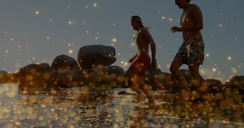 Sunlit duo wading through rocky shoreline at golden hour with warm bokeh glow