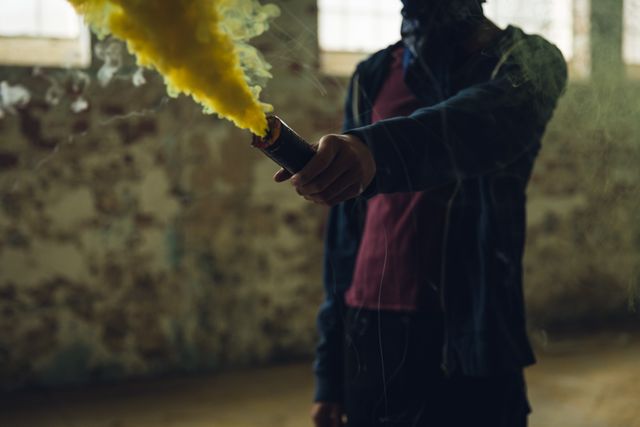 Front view mid section of a hip young biracial man in an empty warehouse, standing and holding in a hand grenade with yellow smoke.